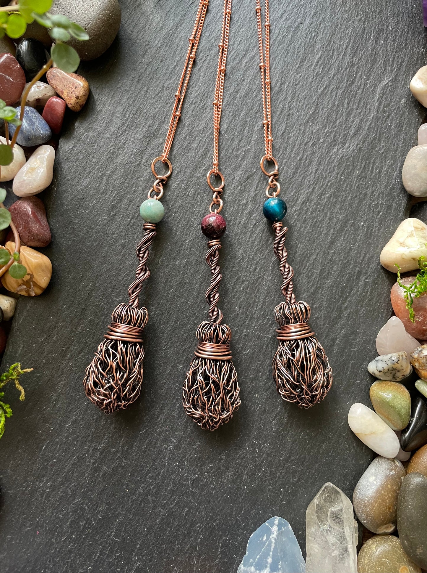 Three copper necklaces with intricate designs on a dark surface with stones and plants.
