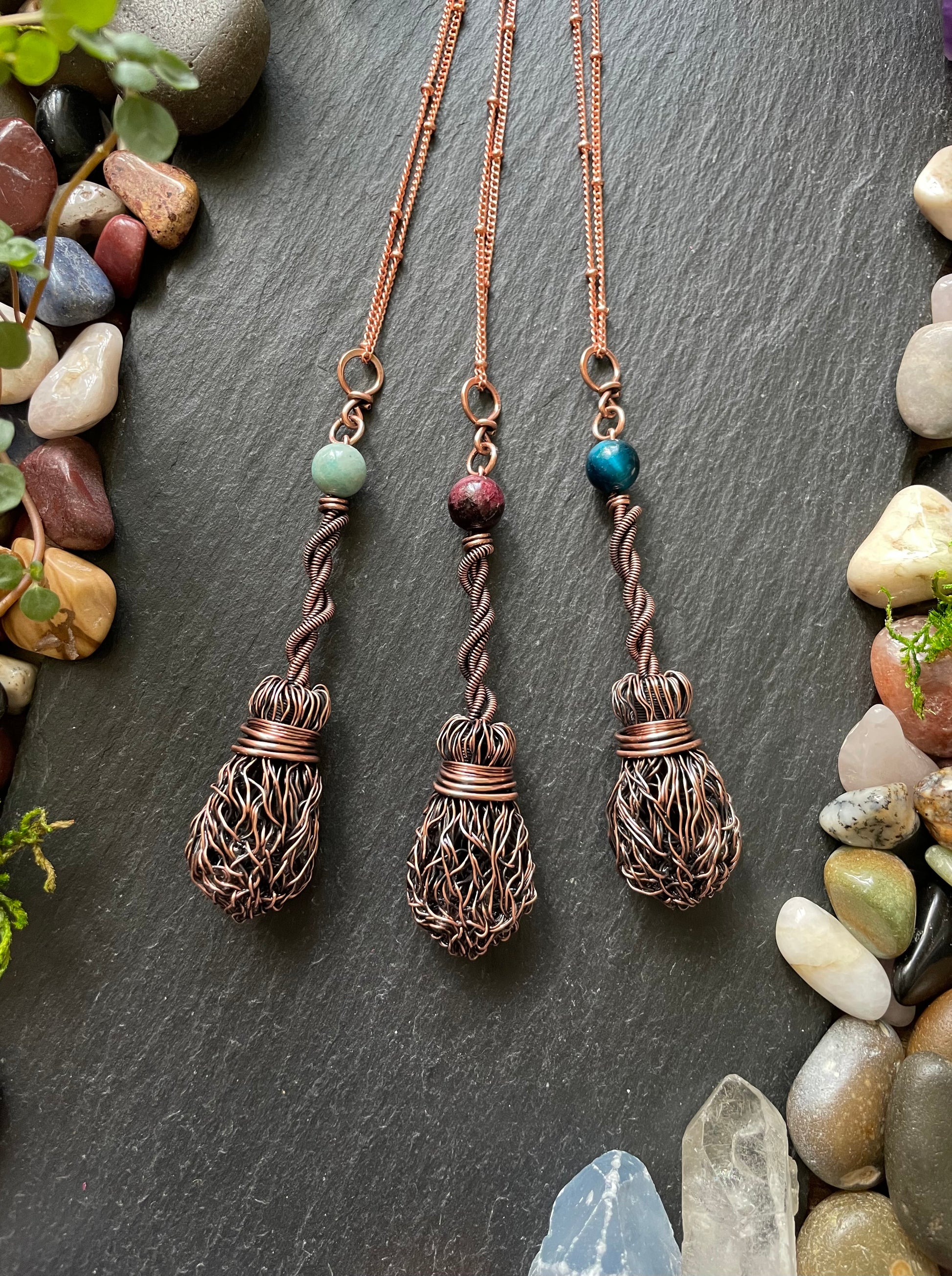Three copper necklaces with intricate designs on a dark surface with stones and plants.