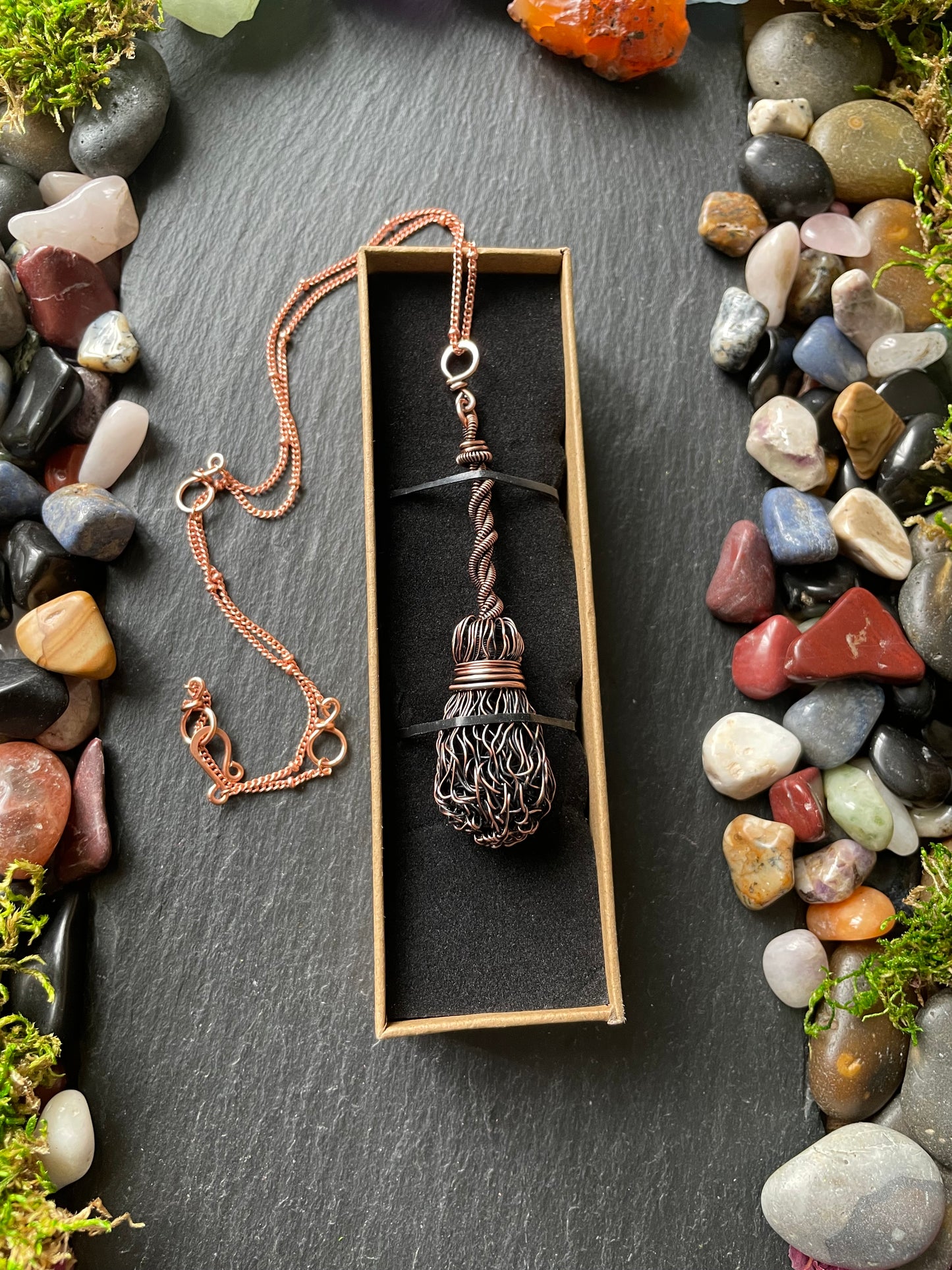 Necklace with a broom pendant in a box on a dark surface with pebbles and moss.