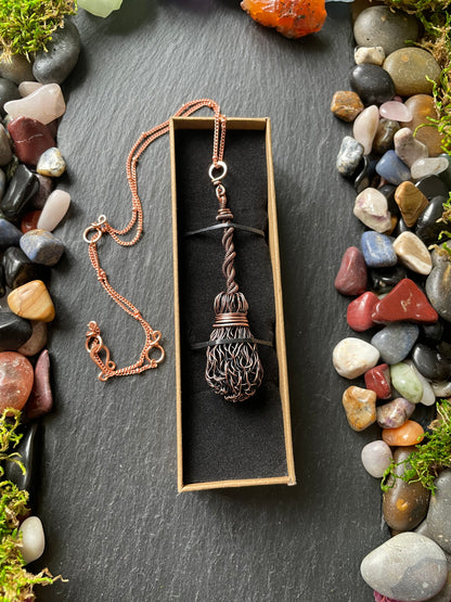 Necklace with a broom pendant in a box on a dark surface with pebbles and moss.