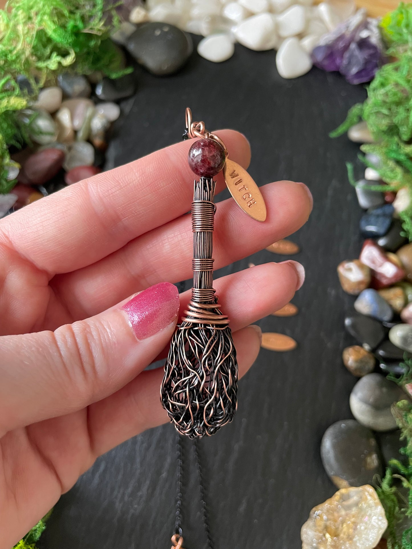 A hand holding a copper broom pendant, set against a dark slate background with decorative stones and moss.
