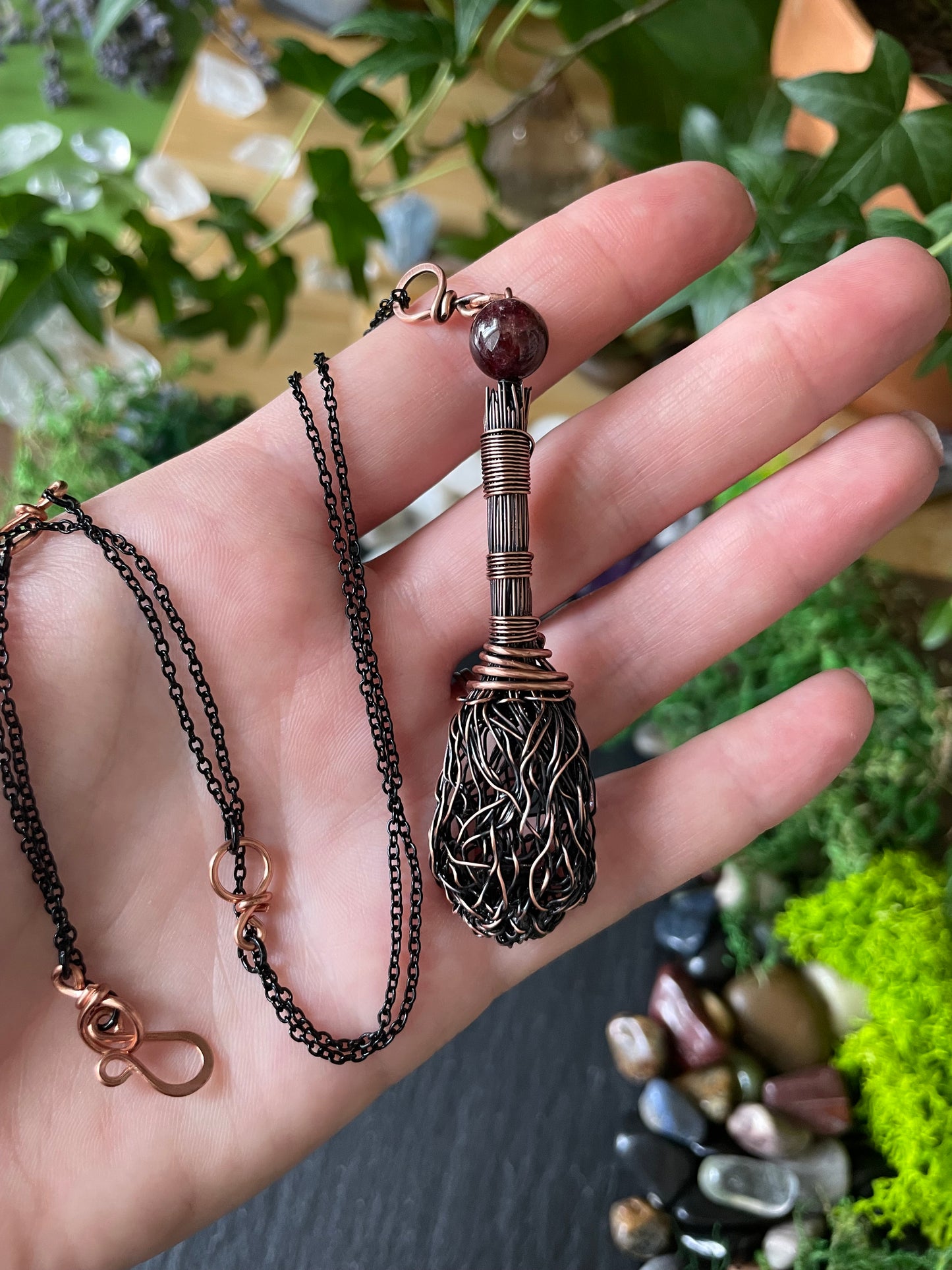 A handmade broom pendant, held in a person's hand against a background of green plants and stones.