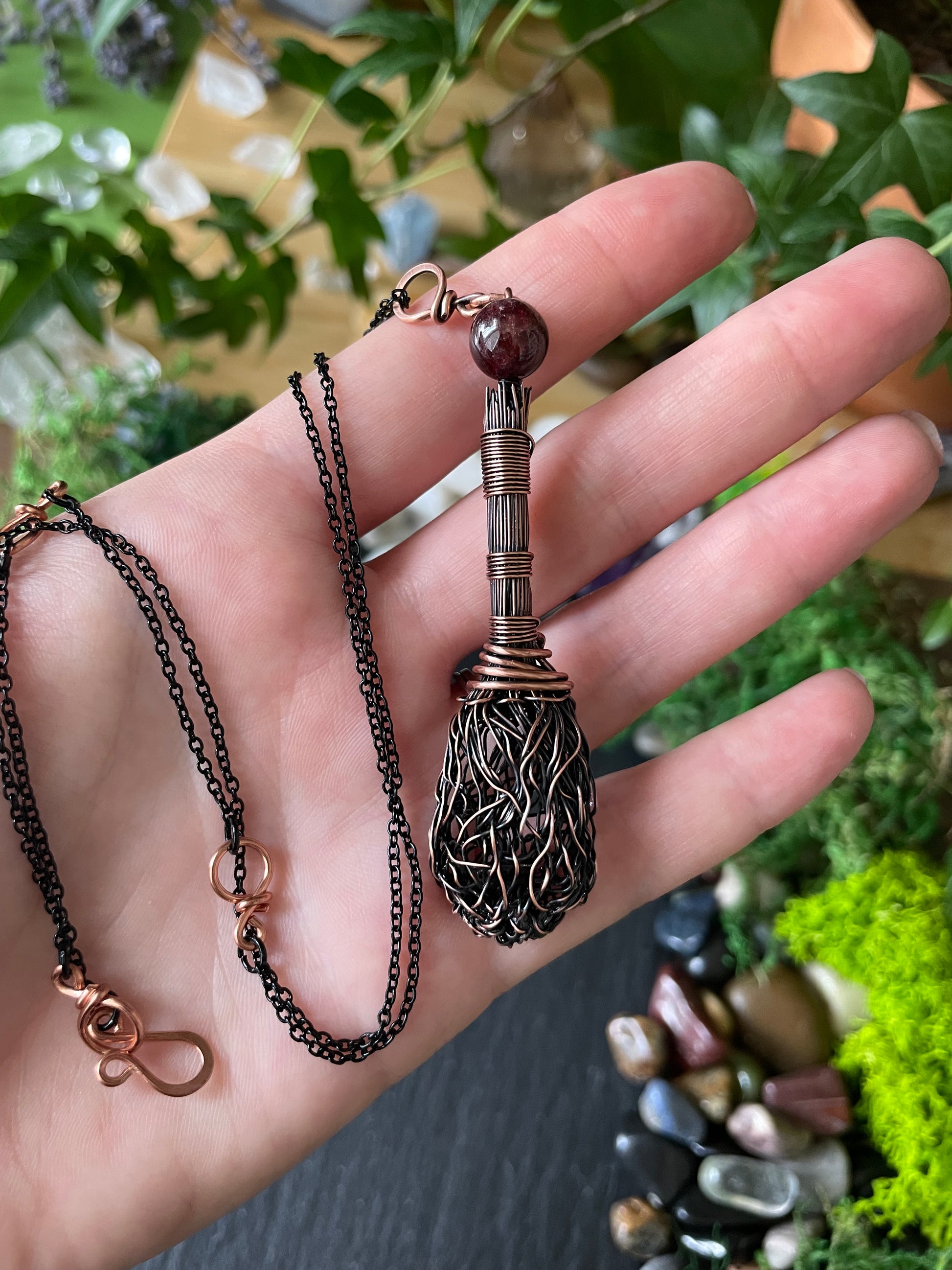 A handmade broom pendant, held in a person's hand against a background of green plants and stones.