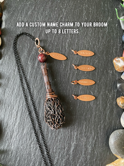 A handmade broom pendant with a red bead, displayed on a dark slate background with four copper name charms and white text overlay.