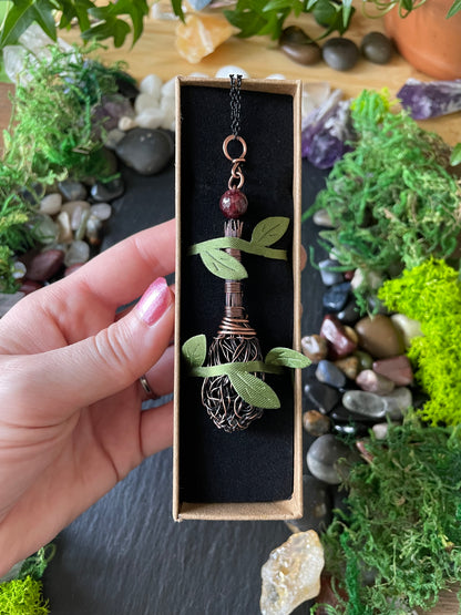 A hand holds a small rectangular black gift box containing a decorative metal broom pendant with a dark red bead, set against a styled background of moss, stones, and crystals.