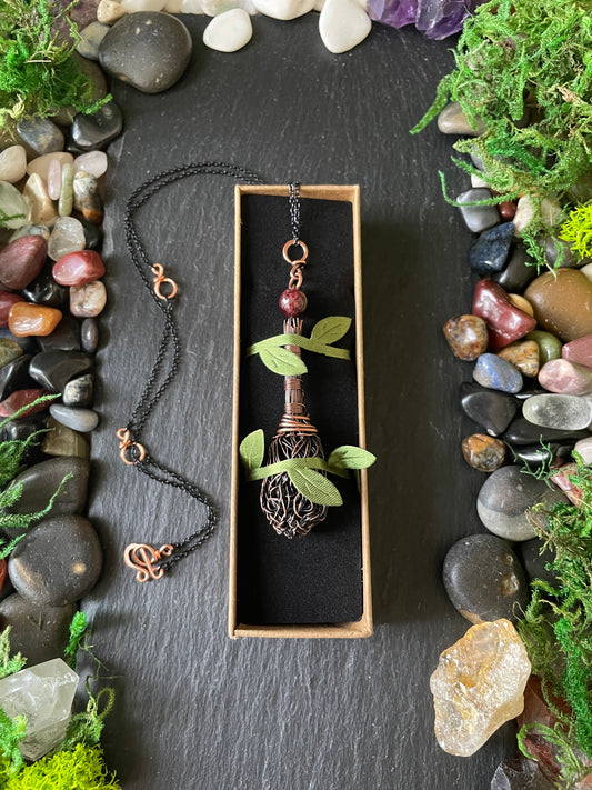 A copper broom pendant necklace displayed in a rectangular box with green leaves and moss, surrounded by decorative river stones and moss on a dark slate background.