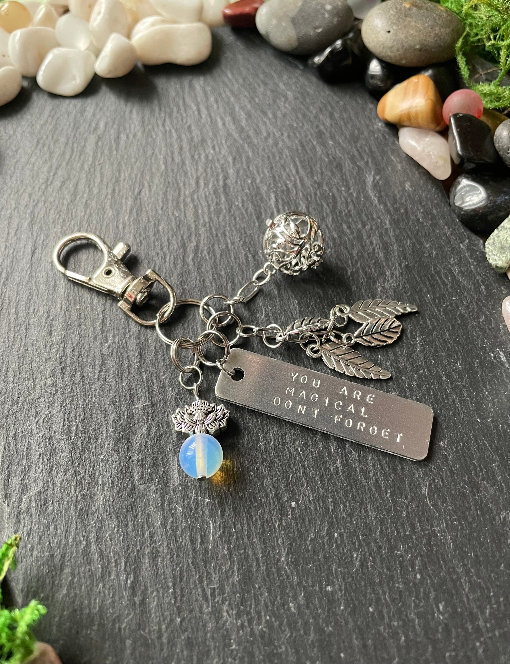 Silver keychain with charms and engraved tag on a stone surface with pebbles and moss.