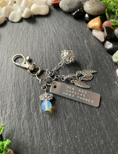 Silver keychain with charms and engraved tag on a stone surface with pebbles and moss.