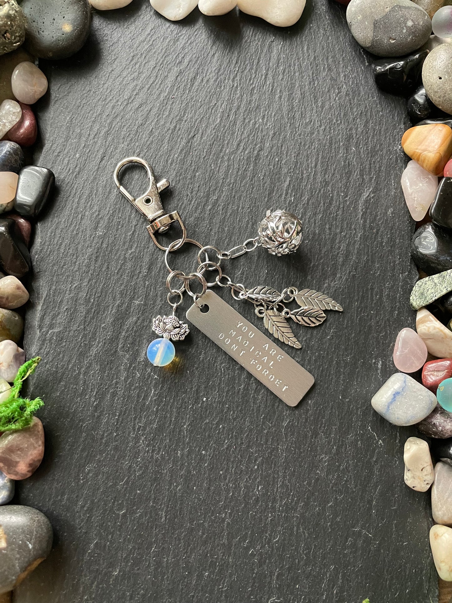 Silver keychain with charms on a dark stone surface surrounded by pebbles.